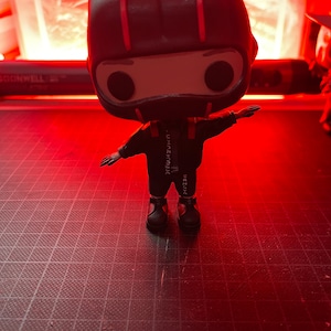 May include: A black and red pop vinyl figure of a person wearing a black jumpsuit with a red stripe down the side and a black helmet with a red stripe. The figure is standing on a black and white grid surface.