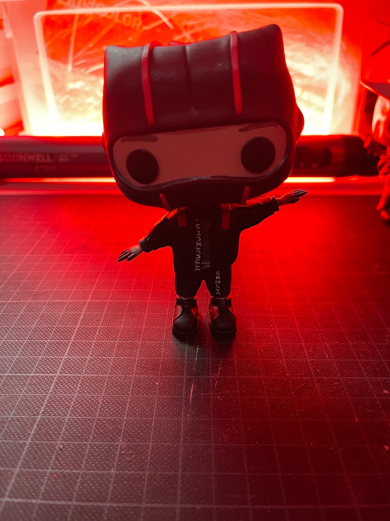 May include: A black and red pop vinyl figure of a person wearing a black jumpsuit with a red stripe down the side and a black helmet with a red stripe. The figure is standing on a black and white grid surface.