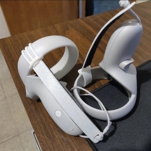 May include: Two white VR controllers with adjustable straps. The controllers have a white strap with a textured surface and a white strap with a black strap.
