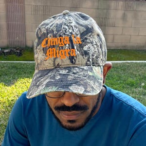 May include: Camouflage baseball cap with the text "Chinga la Migra" in orange script. The cap has a curved brim and a classic six-panel design. The hat is made of a durable fabric and features a camouflage pattern in shades of green, brown, and gray.