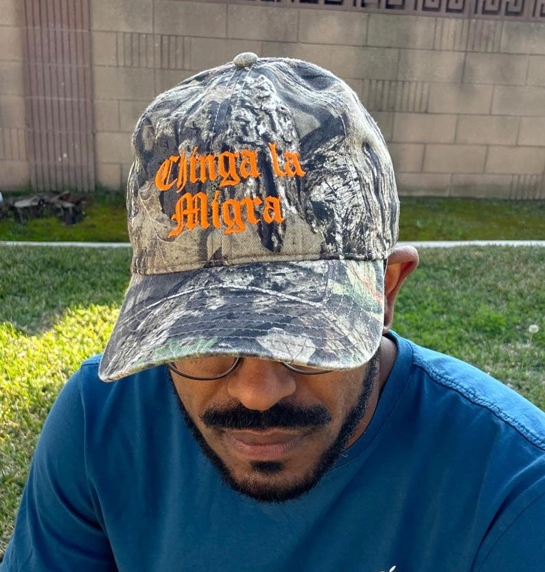 May include: Camouflage baseball cap with the text "Chinga la Migra" in orange script. The cap has a curved brim and a classic six-panel design. The hat is made of a durable fabric and features a camouflage pattern in shades of green, brown, and gray.