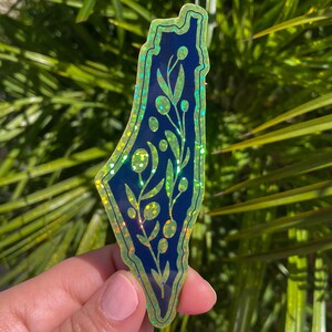 K&ouml;nnte beinhalten: Ein gr&uuml;ner und blauer, schimmernder Aufkleber mit der Umrissform Israels und einem floralen Design im Inneren. Der Aufkleber wird in einer Hand vor einem gr&uuml;nen, bl&auml;ttrigen Hintergrund gehalten.
