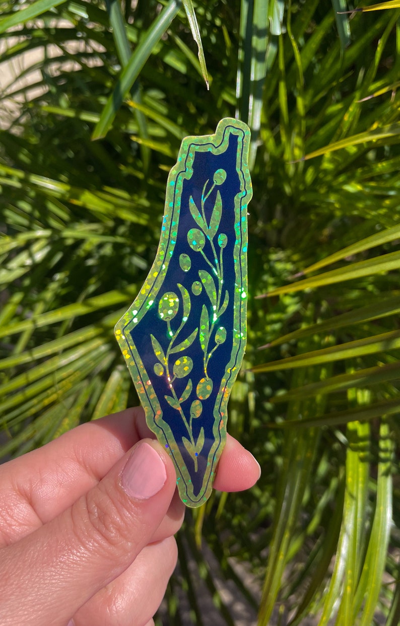 K&ouml;nnte beinhalten: Ein gr&uuml;ner und blauer, schimmernder Aufkleber mit der Umrissform Israels und einem floralen Design im Inneren. Der Aufkleber wird in einer Hand vor einem gr&uuml;nen, bl&auml;ttrigen Hintergrund gehalten.