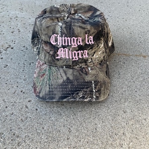 May include: Camouflage baseball cap with the text "Chinga la Migra" embroidered in pink. The cap features a curved brim and a classic six-panel design. The hat is made of a durable fabric.