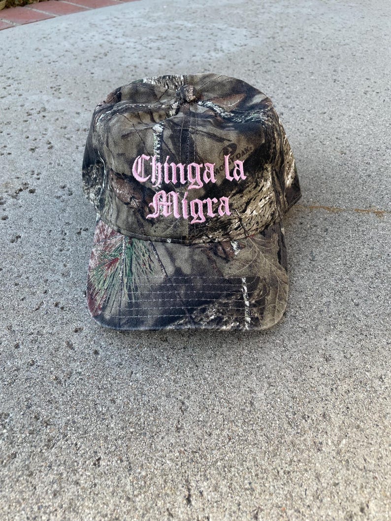 May include: Camouflage baseball cap with the text "Chinga la Migra" embroidered in pink. The cap features a curved brim and a classic six-panel design. The hat is made of a durable fabric.