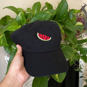 Puede incluir: Una gorra de béisbol negra con una rodaja de sandía roja y verde bordada en la parte delantera.