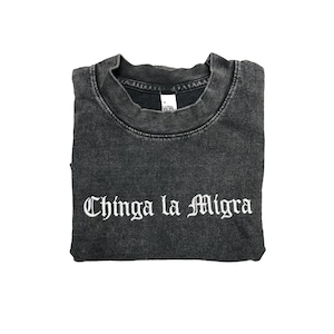May include: Folded charcoal gray t-shirt with the phrase "Chinga la Migra" in white gothic lettering. The shirt has a crew neck and a slightly faded appearance, suggesting a vintage or distressed style.