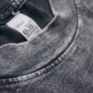 Może przedstawiać: Zbliżenie na szary t-shirt z białą metką z napisem "6.5 OUNCE" i "REPRELLE"