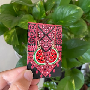 May include: A pair of gold watermelon slice earrings on a black and red patterned card. The earrings are made of metal and have a red, green and white watermelon slice design.