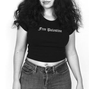 以下が含まれることがあります： ゴシック体で「Free Palestine」と書かれた黒のクロップドトップス。トップスは青いデニムジーンズと組み合わせています。画像は白黒です。