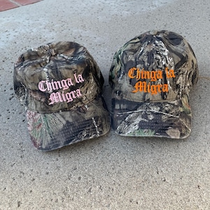 May include: Two camouflage baseball caps with the text "Chinga la Migra" embroidered on the front. One hat has pink lettering, and the other has orange lettering. The hats are made of a camouflage fabric.