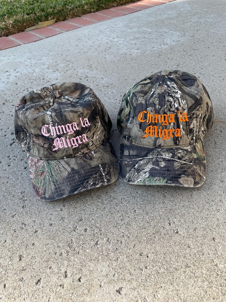 May include: Two camouflage baseball caps with the text "Chinga la Migra" embroidered on the front. One hat has pink lettering, and the other has orange lettering. The hats are made of a camouflage fabric.