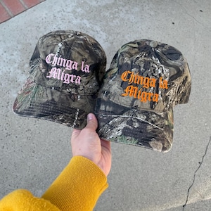 May include: Two camouflage baseball caps with the text "Chinga la Migra" embroidered in pink and orange. The caps are held up by a person wearing a yellow sweater.