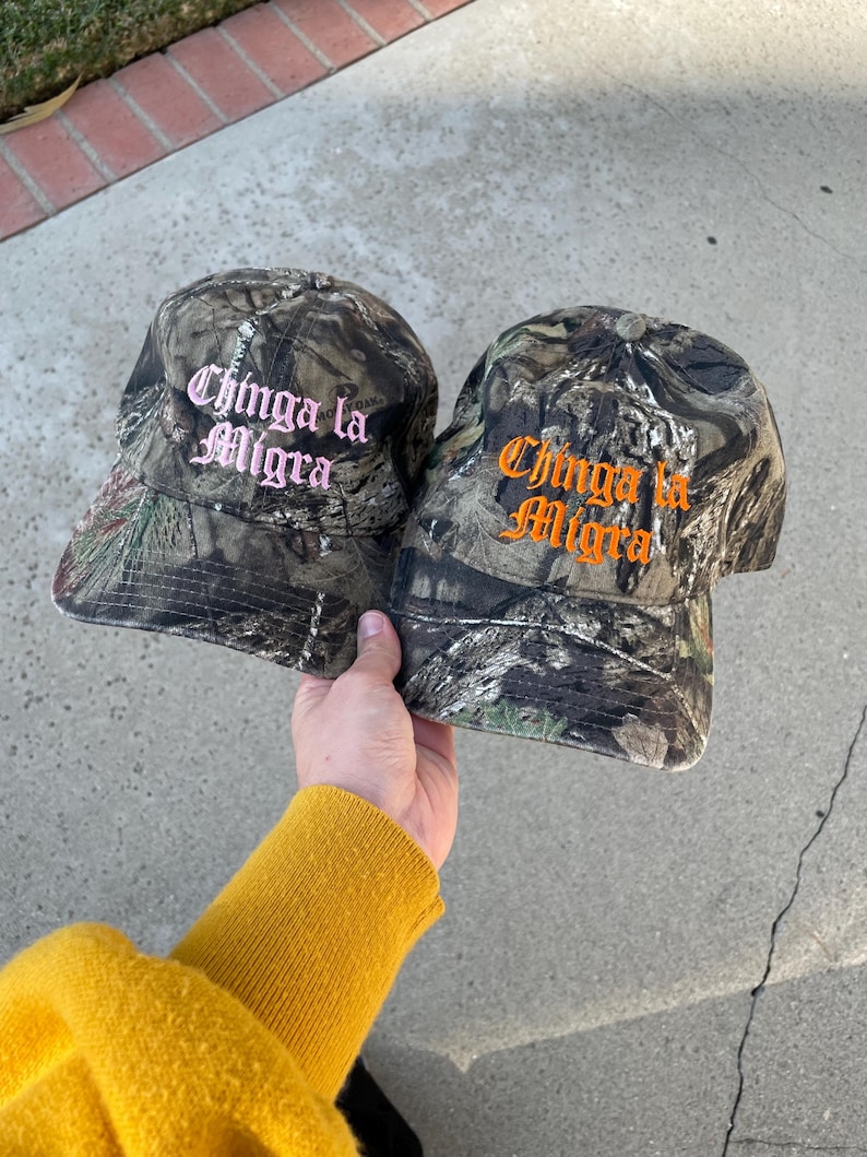 May include: Two camouflage baseball caps with the text "Chinga la Migra" embroidered in pink and orange. The caps are held up by a person wearing a yellow sweater.