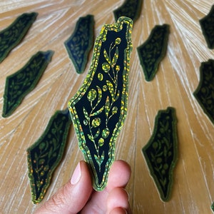 K&ouml;nnte beinhalten: Ein schwarzer und gr&uuml;ner Glitzeraufkleber mit der Umrissform Israels und einem floralen Design im Inneren.