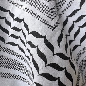 May include: White and black keffiyeh scarf with a geometric pattern. The scarf features a repeating design of dotted lines, teardrop shapes, and floral motifs. The fabric is folded, showing the intricate details of the design.