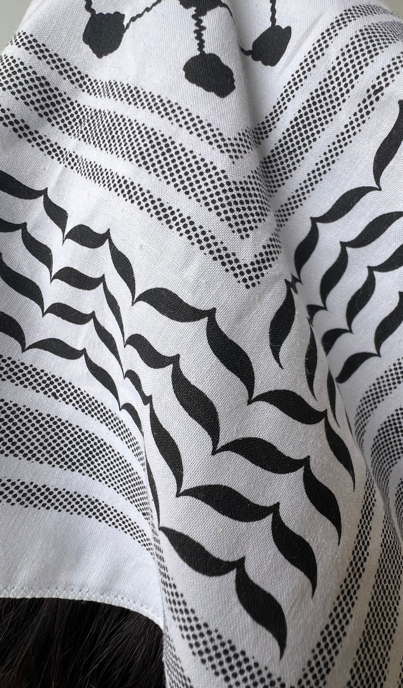 May include: White and black keffiyeh scarf with a geometric pattern. The scarf features a repeating design of dotted lines, teardrop shapes, and floral motifs. The fabric is folded, showing the intricate details of the design.