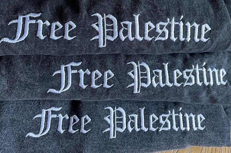 Może przedstawiać: Trzy szare koszulki ułożone jedna na drugiej. Każda koszulka ma haftowany napis "Free Palestine" w kolorze białym z przodu.