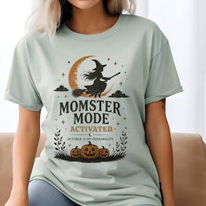 May include: A light sage green t-shirt featuring a graphic of a witch flying on a broomstick in front of a crescent moon. Below, the text reads MOMSTER MODE ACTIVATED. OCTOBER IS MY PERSONALITY. Three jack-o'-lanterns sit at the bottom of the design.