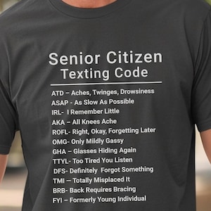May include: Dark gray t-shirt with the text "Senior Citizen Texting Code" in white. The shirt lists humorous acronyms and their meanings, such as "ATD - Aches, Twinges, Drowsiness" and "FYI - Formerly Young Individual."
