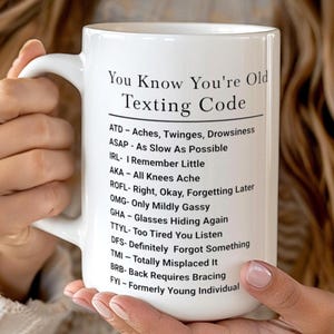 May include: White ceramic mug with the text "You Know You're Old Texting Code" and a list of acronyms and their meanings. The mug is held by a person.