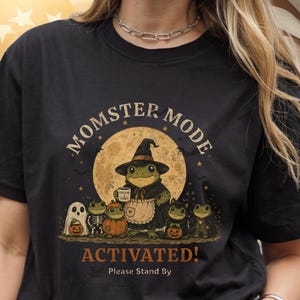 May include: A black t-shirt featuring a graphic of a frog dressed as a witch holding a mug that says CAFFEINE & CHAOS, surrounded by smaller frogs in Halloween costumes. The text reads MOMSTER MODE ACTIVATED! Please Stand By.