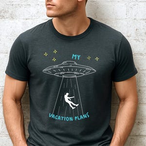 May include: Dark gray t-shirt with a white graphic of a UFO beaming up a person. The text "MY VACATION PLANS" is in light blue below the UFO. Four yellow stars are above the UFO.