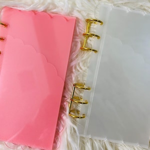 May include: Two planner covers with gold rings. One is a translucent pink, the other is clear. Both have a scalloped edge and are open, revealing the interior. The covers are on a white, fluffy surface.