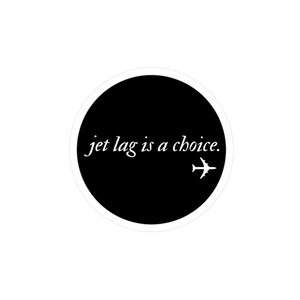 以下が含まれることがあります： 黒い背景と白い縁取りの丸いステッカー。「jet lag is a choice」というテキストが白いスクリプトで書かれています。白い飛行機のグラフィックがテキストの右側にあります。