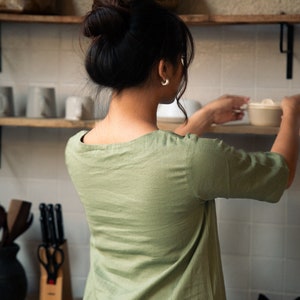 Puede incluir: Una mujer con una camisa verde salvia de manga corta y cuello redondo. Est&aacute; de pie en una cocina y alcanza un taz&oacute;n de cer&aacute;mica blanco en un estante.