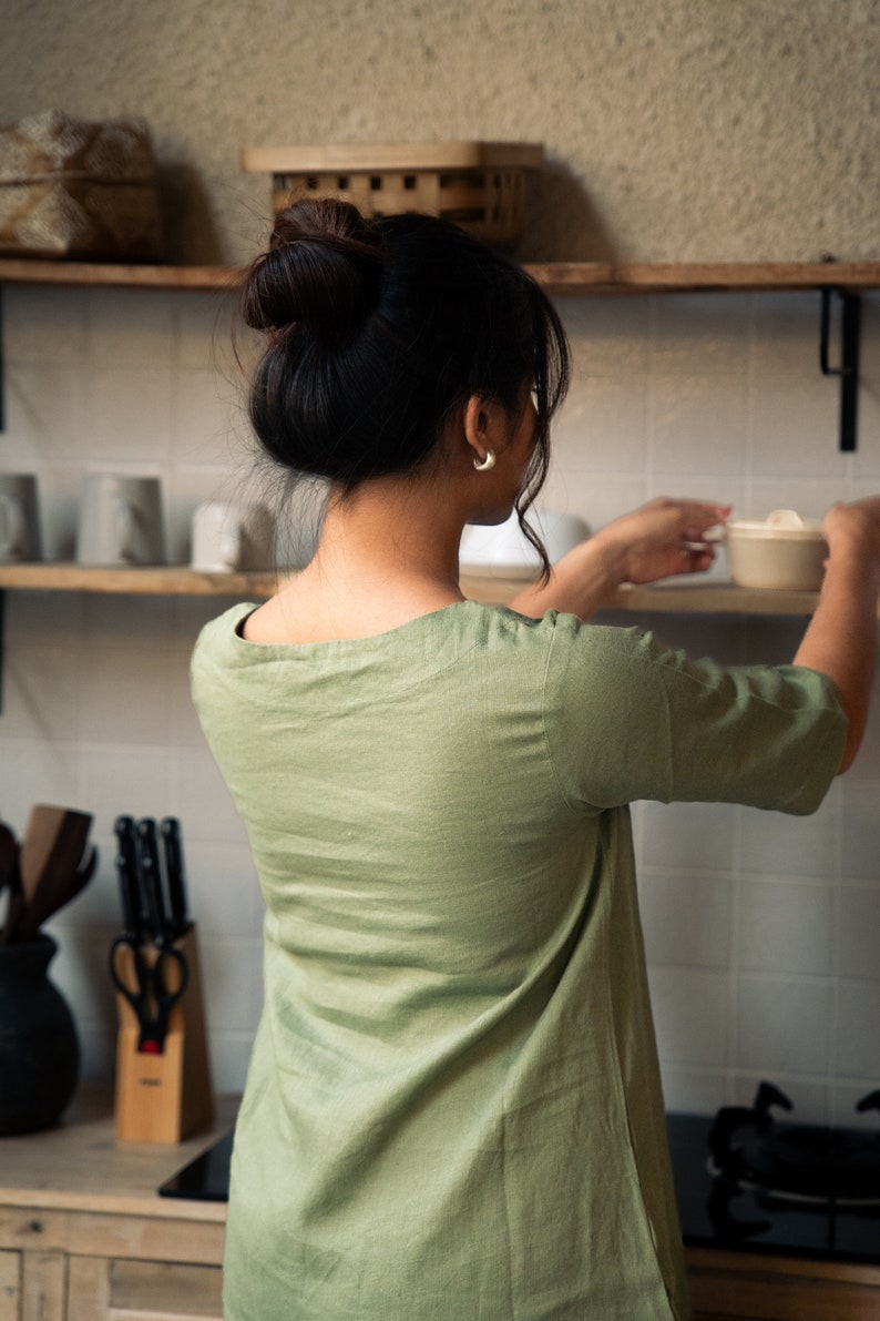 Puede incluir: Una mujer con una camisa verde salvia de manga corta y cuello redondo. Est&aacute; de pie en una cocina y alcanza un taz&oacute;n de cer&aacute;mica blanco en un estante.