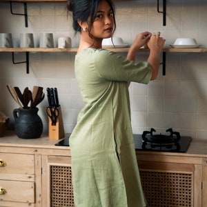 Puede incluir: Una mujer con un vestido de lino verde salvia est&aacute; de pie en una cocina. Est&aacute; alcanzando un estante para tomar una taza de cer&aacute;mica blanca.