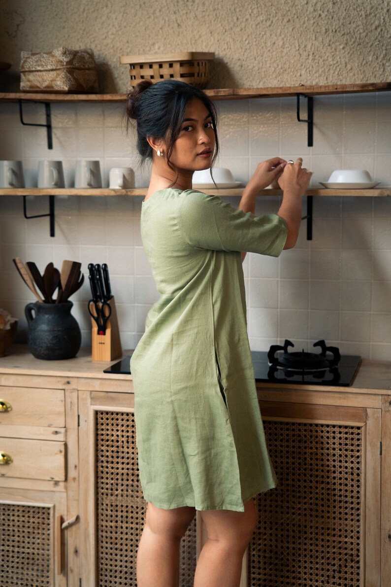 Puede incluir: Una mujer con un vestido de lino verde salvia est&aacute; de pie en una cocina. Est&aacute; alcanzando un estante para tomar una taza de cer&aacute;mica blanca.