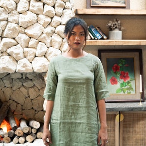 Puede incluir: Una mujer con un vestido de lino verde salvia de manga corta y cuello redondo. Est&aacute; de pie frente a una pared de piedra con una chimenea y una estanter&iacute;a con cuadros enmarcados y libros.