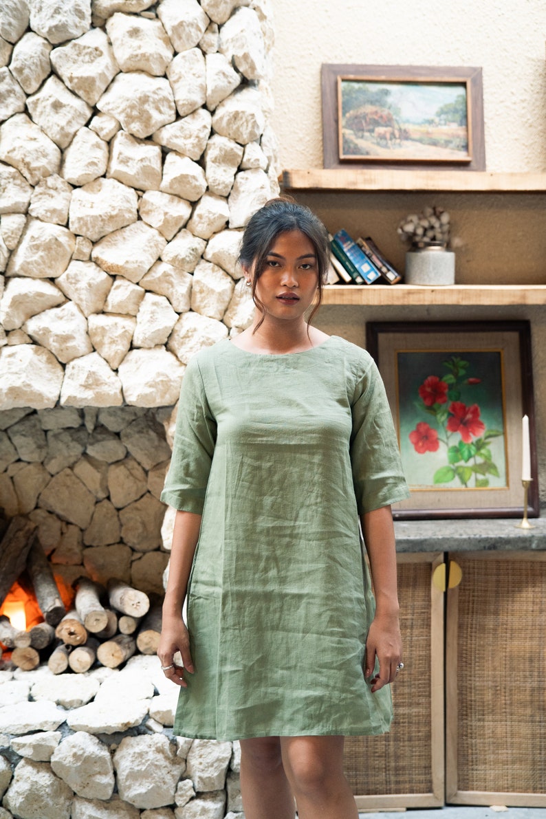 Puede incluir: Una mujer con un vestido de lino verde salvia de manga corta y cuello redondo. Est&aacute; de pie frente a una pared de piedra con una chimenea y una estanter&iacute;a con cuadros enmarcados y libros.