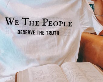 May include: White t-shirt with the text "We The People Deserve The Truth" in black. The shirt is being held up, with an open book and a glass of water in the foreground.