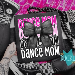 May include: Black t-shirt with the words "DANCE MOM" in white and pink, featuring a large bow graphic. The image includes black Converse sneakers, a black beaded necklace, and a checkered hair clip.