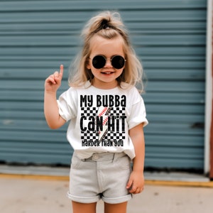 May include: A white t-shirt with a black and white checkered design featuring a baseball bat and ball with a lightning bolt. The text on the shirt reads "My Bubba Can Hit Harder Than You".