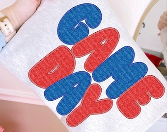 Op de afbeelding: Een lichtgrijs kussen met de tekst "GAME DAY" in grote, vette letters. De letters zijn rood omlijnd en blauw gevuld, wat een opvallend contrast creëert. Het kussen wordt door een persoon vastgehouden.