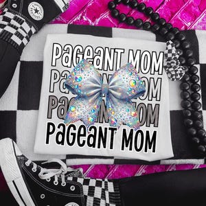 May include: White t-shirt with the text "Pageant Mom" in a stacked design. A large, silver bow with iridescent rhinestones is centered on the shirt. The image is set against a black and white checkered pattern.