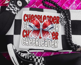 Puede incluir: Camiseta blanca con la palabra "Cheerleader" repetida cuatro veces en texto rojo y blanco. Un lazo brillante rojo y blanco está centrado en la camiseta.