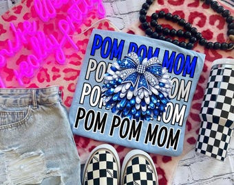 Può includere: Una maglietta blu chiaro con il testo "Pom Pom Mom" ripetuto quattro volte. Un grande pompon blu e bianco con un fiocco argentato è centrato sulla maglietta.