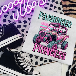 May include: White t-shirt with a pink and green graphic of an off-road vehicle with the text "Passenger Princess" on it.