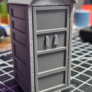 May include: A gray 3D printed model of a small building with a rounded roof and a door with two small rectangular cutouts. The building has a textured surface and appears to be a miniature model of a public restroom.