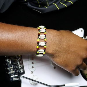 May include: A beaded bracelet featuring white seashells and colorful beads in red, yellow, and green. The bracelet is worn on a wrist, with a black background and a guitar visible in the background.
