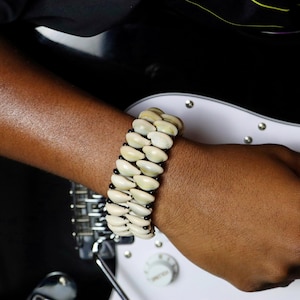 May include: A cream-colored shell bracelet with black beads. The bracelet is made of multiple rows of shells and is worn on a wrist. The background includes a black surface and a white guitar.