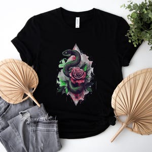 May include: Black t-shirt with a graphic of a green snake and a pink rose. The design is set against a geometric shape with watercolour accents. The shirt is paired with grey jeans and a decorative fan.