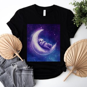 Puede incluir: Camiseta negra con un gráfico de un perezoso morado durmiendo en una luna creciente contra un cielo estrellado. El diseño está centrado en la camiseta. La camiseta se combina con vaqueros grises y dos abanicos beige.