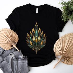 May include: Black t-shirt featuring a geometric design in shades of teal, gold, and yellow. The design is centered on the shirt. The shirt is paired with gray jeans and two natural woven fans. A small green plant is in the background.
