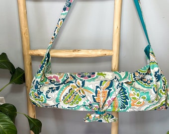 May include: A teal and white paisley patterned yoga mat carrier with a long strap. The strap is tied in a bow at the front of the carrier.